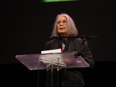 Anne Carson står på en scene ved en klar talerstol med en mikrofon foran en mørk baggrund.